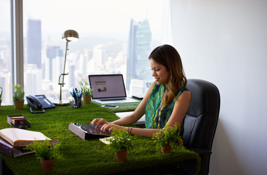 Concept of ecology and environment: Young business woman working in modern office with table covered of grass and plants. She types on tablet pc